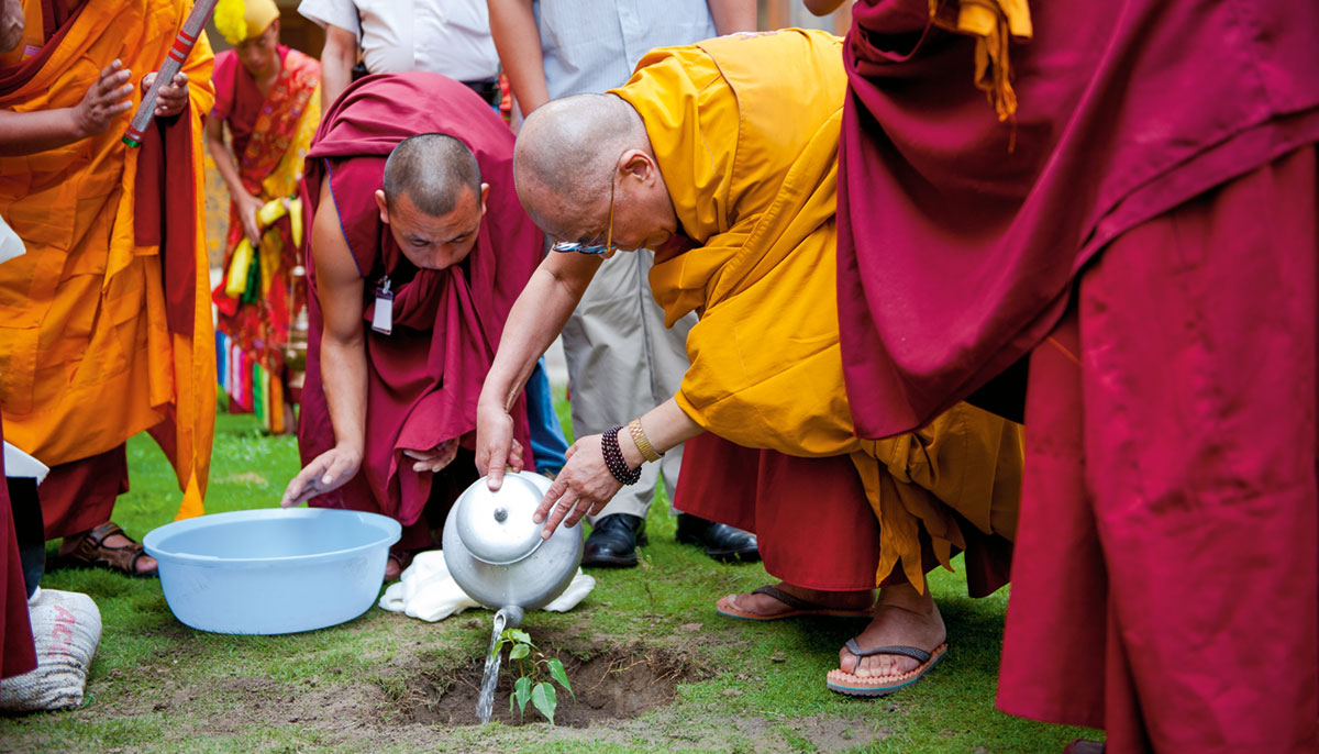 The Dalai Lama pouring water from a teapot into a hole in the ground. He is wearing robes and surrounded by other people in robes.