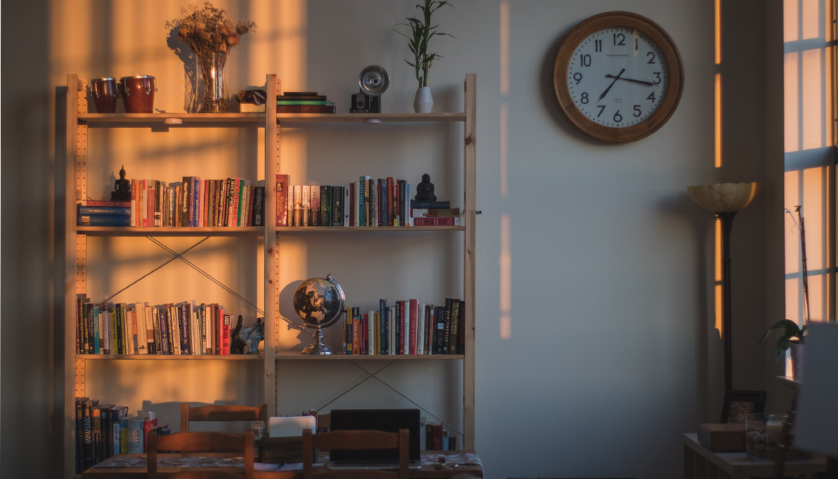 A photo of a bookshelf covered in books with sunset-coloured lighting overtop of it. There is a clock to the right of the bookshelf.