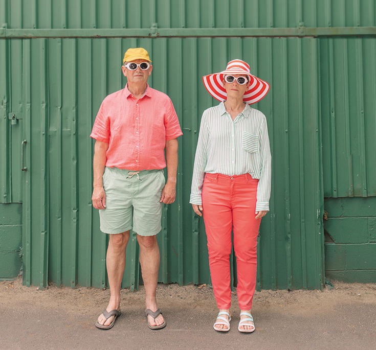Two people stand in summer clothes. The man is wearing a yellow hat with a pink shirt and green shorts. The woman is wearing coral pants and a long sleeve stripe shirt and striped hat.