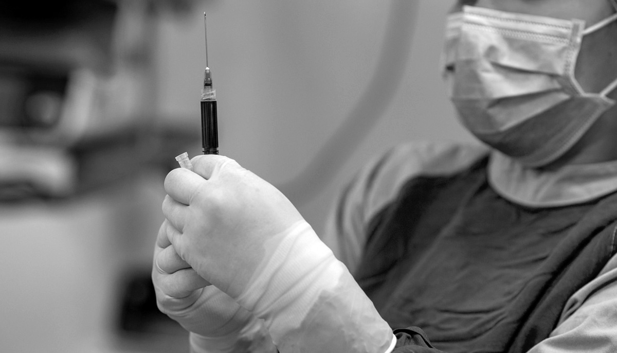 Black and white photo of someone wearing gloves and a mask, holding a vaccine.