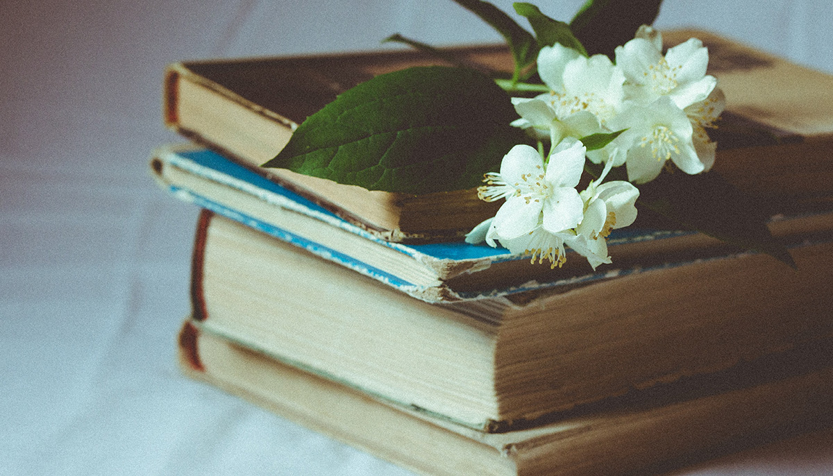 A pile of four aged books with flowers on top of the stack.