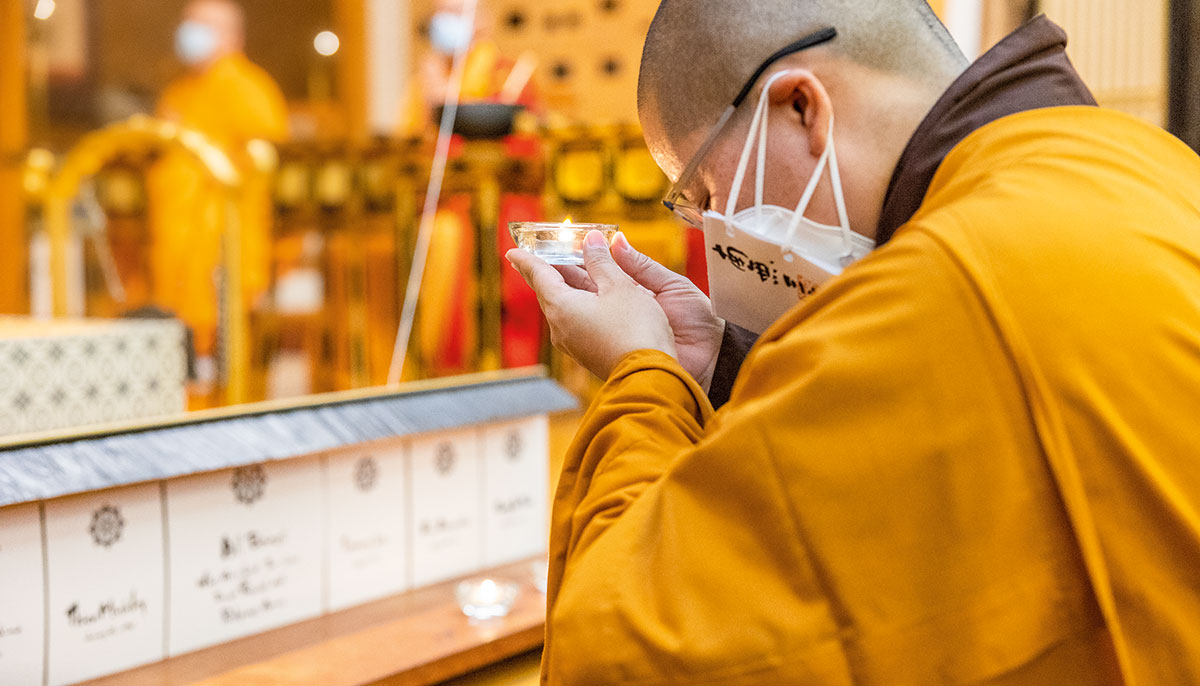 A man in orange robes holds up a candle. He is wearing a surgical mask and the photo is taken from profile view.