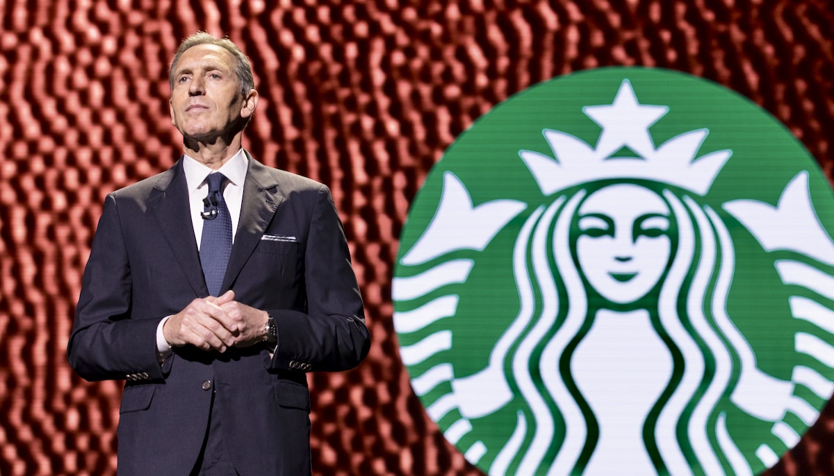 Howard Schultz stands on a stage in front of a Starbucks logo.