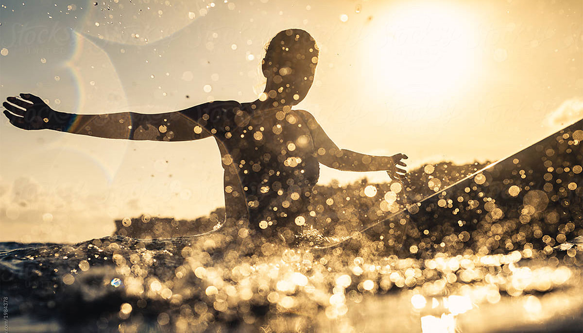 Woman on surfboard with arms outstretched and sun in background.