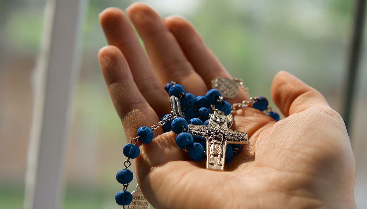 A hand holds a blue Catholic rosary.