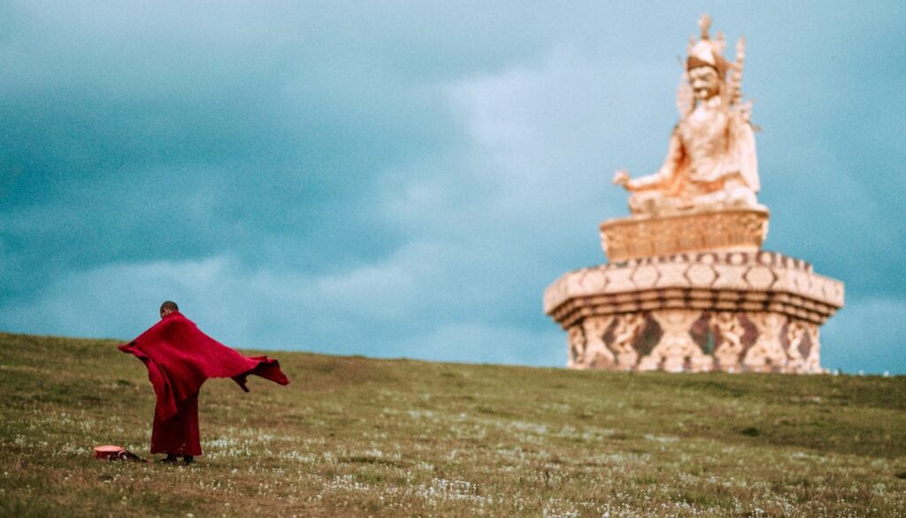 A monk standing in a field, with a statue of Padmasambhava in the distance.