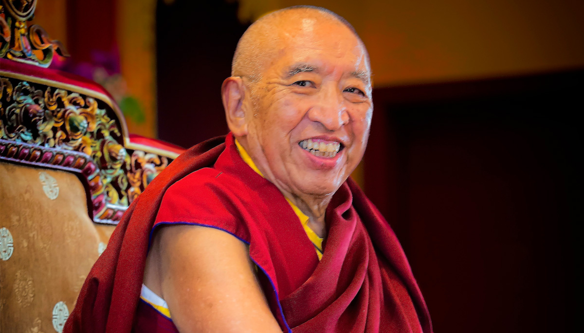 Thrangu Rinpoche smiles, wearing red robes and seated in a chair