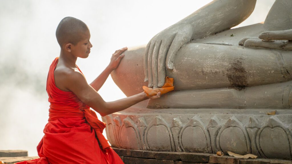 A novice monk uses a cloth to clean a large Buddha statue.