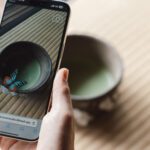 A hand holds a smartphone displaying a photo of a matcha bowl with a digital butterfly overlay. The real bowl, filled with green tea, sits on a tatami mat.